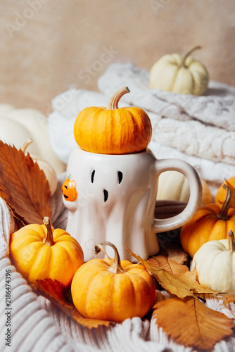 Whimsical ghost-shaped mug topped with a pumpkin, surrounded by miniature pumpkins, autumn leaves, and cozy knit fabric in warm seasonal tones.