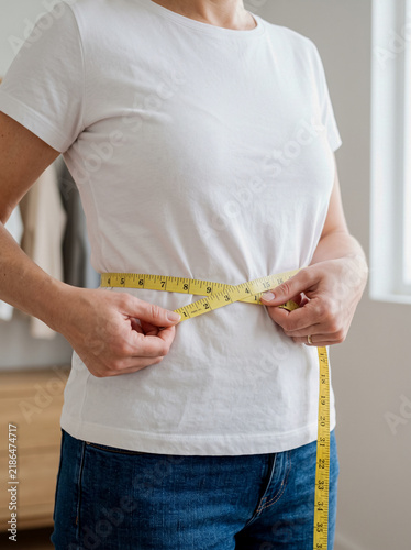 Close-up of a woman in a white t-shirt measuring her waist with a yellow measuring tape.
