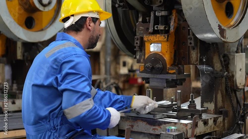 Male adult industrial worker operating metal stamping press machine, wearing protective gear inside busy factory manufacturing facility, focused precision manufacturing, workplace safety