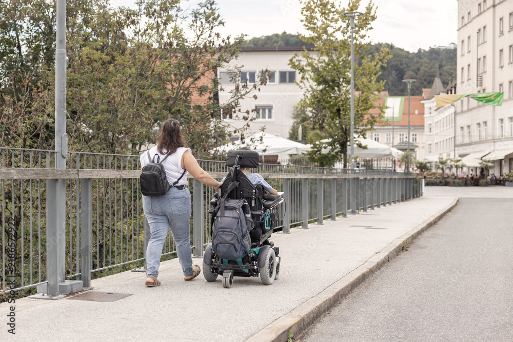 Obraz premium Powered wheelchair user moving along an outdoor walkway accompanied by a Personal assistant, supporting independent mobility.