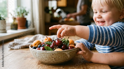 Toddler's Fruitful Discovery: A beaming toddler reaches for a juicy cherry from a brimming bowl of fresh fruit, experiencing the simple joy of healthy eating and discovery in a home kitchen setting.