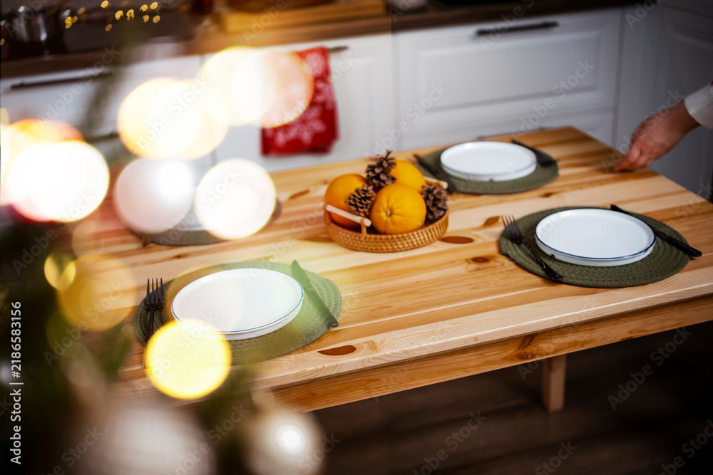 Naklejka premium Christmas wooden table setting with empty plates and fresh oranges — holiday dinner preparation. Festive home kitchen interior featuring warm fairy lights. Cozy winter evening meal anticipation.