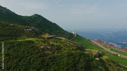 Expansive aerial panorama showcasing a historic red-and-white lighthouse nestled on a verdant mountainside, overlooking the vast, serene Atlantic Ocean and a winding coastal road, capturing the