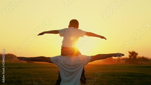 Dad plays with son, beloved child sits on father shoulders with arms raise, sun sky. Happy family plays park. Dad plays outdoors with child, dream traveling by plane. Dad child superhero fly, sunset
