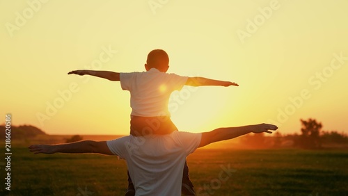 Dad plays with son, beloved child sits on father shoulders with arms raise, sun sky. Happy family plays park. Dad plays outdoors with child, dream traveling by plane. Dad child superhero fly, sunset
