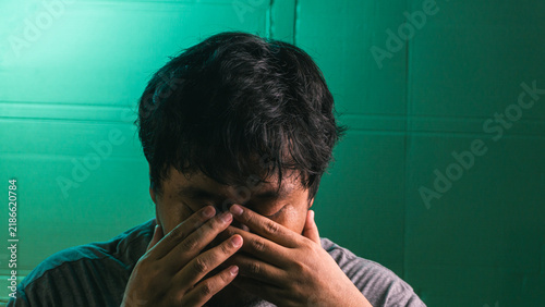 Foto A stressed young indonesian male bows the head and pinches the bridge of the nose, demonstrating severe frustration, a migraine, or mental fatigue against a dark green textured wall
