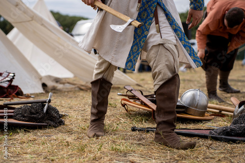 Konstfotografi Historical reenactor in medieval tunic and leather boots holding axe standing outdoors near armor equipment and white tent camp