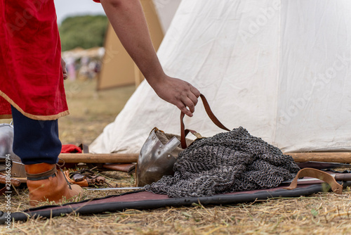 Konstfotografi Person reaching down to lift metallic chainmail armor mesh from ground near white historical tent during reenactment camp