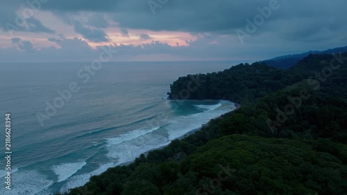 Sunset Over Rugged Coast With Flowing Ocean Waves And Forested Cliffs. Aerial Footage Showing Vibrant Sunset Illuminating Rocky Shoreline Alongside Crashing Sea Waves