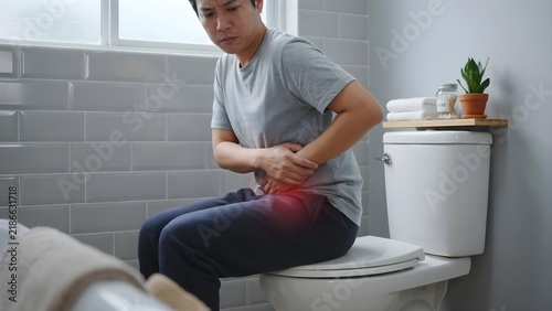 Young Asian woman sitting on the toilet bowl with tissue paper on hand and suffering from constipation, diarrhea, stomach ache or cramps. Copy space