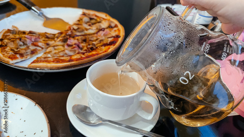 A hand pours tea into a white cup. A plate of pizza is visible in the background. The setting is casual dining with a focus on food and beverages.