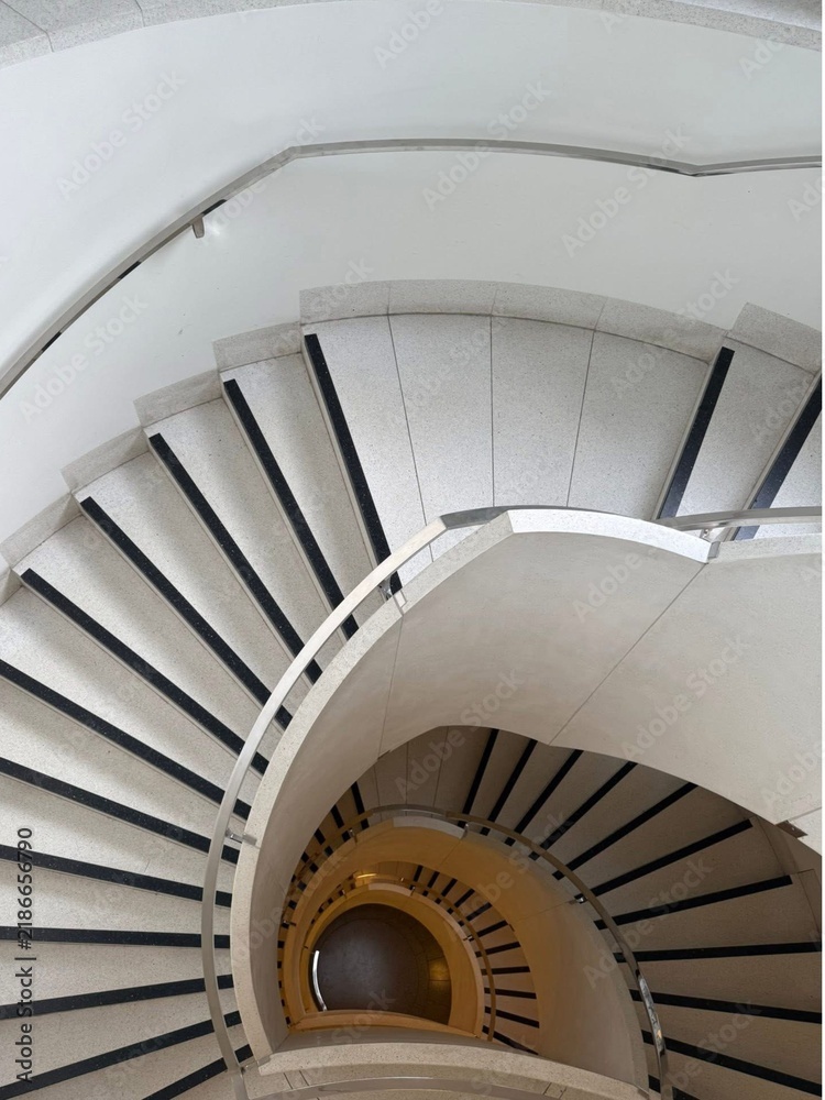 Fototapeta premium Looking down at a grand modern spiral staircase with white steps and black safety strips, creating a beautiful geometric pattern.