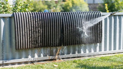 Black ribbed rubber mat being cleaned with high pressure washer on corrugated metal fence outdoors during bright sunny day.