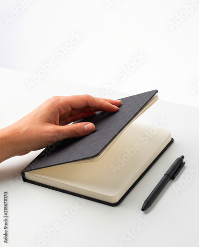 Hand Opening Black Hardcover Notebook with Pen on White Desk Mockup
