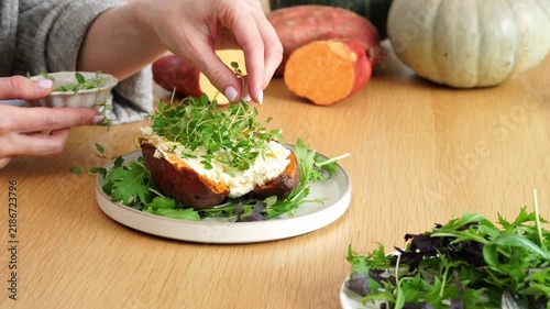 Close-up hands garnishing hollowed sweet potato with fresh greens on wooden table in warm autumn light on rustic kitchen table, white plate, autumn vegetables background