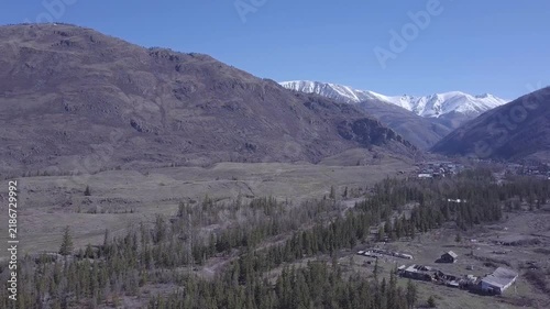Mountain valley landscape stretches beneath snow covered peaks as aerial perspective reveals forest patches and scattered buildings. Broad ridges frame open terrain under clear blue sky.