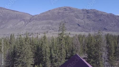 Wooden cabins overlook forested slopes in mountain resort, while rugged mountains rise beyond tall evergreens. Log houses, red roofs, and open alpine landscape create peaceful holiday setting.