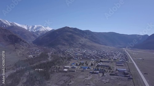 Aerial drone flight surveys town spread across mountain valley, following roads past clustered buildings and open ground. Remote settlement rests beneath rugged slopes and distant snow covered peaks.