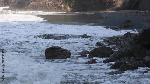 Waves crash against dark, jagged rocks at the shore. White foam spills over rugged stones with force and energy. Rocks dot the beach, some wet, some dry. Sandy ground meets sea spray