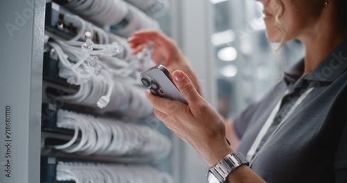 Female Network Technician Performs Physical Port Audit Beside Server Rack, Using Smartphone to Verify Cable Identification and Network Records. Concept of Maintains Data Center Infrastructure.