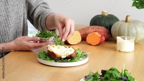 Hands placing fresh thyme herb garnish onto openfaced baked sweet potato on rustic autumn table with pumpkin and squash in warm kitchen light, close up food preparation scene