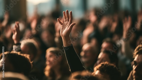 A raised hand stands out among a crowd in a dark conference hall. This photo represents participation, asking questions, and voting.
