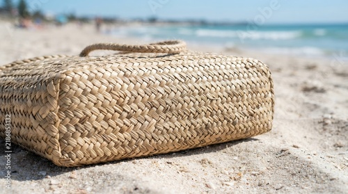 Woven Straw Beach Bag Resting on Sandy Shoreline with Ocean Waves in Background.
