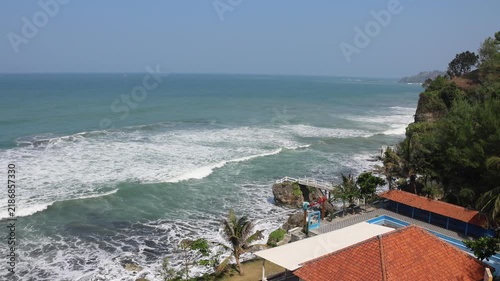 Waves crash against rocks near a coastal property. Green grass and palm trees frame the scene. Small building sits near the shore. Sunlight glints off the moving water