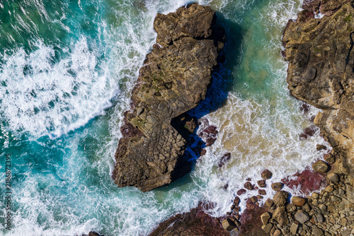 Description: Downward aerial view of ocean waves crashing onto rugged rock formations along the Yamba coastline, creating white foam and natural patterns beside the Pacific Ocean.