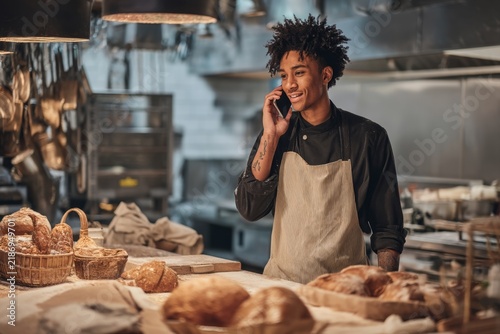 Smiling young baker on phone in modern bakery surrounded by fresh artisan bread and pastry products showcasing a vibrant culinary scene. Generative AI