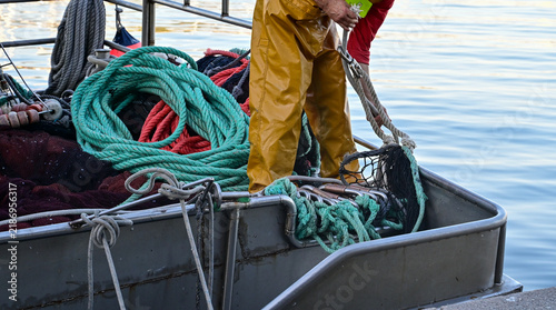 A professional fisherman in yellow waterproof clothing works aboard a small fishing boat, handling ropes and fishing nets beside calm coastal waters. 
