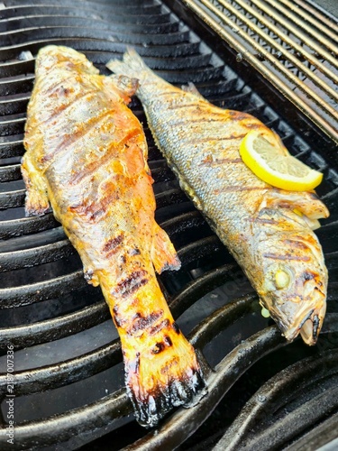 Two whole fish cooking on a hot outdoor barbecue grill with visible grill marks, smoke and fresh lemon, authentic summer seafood preparation.