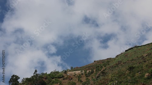 Blue sky above green hillside. Soft clouds drift lazily overhead. Vegetation covers slopes gently. Meadows and trees mix naturally. Scene feels calm and serene. Ideal for nature or travel backgrounds