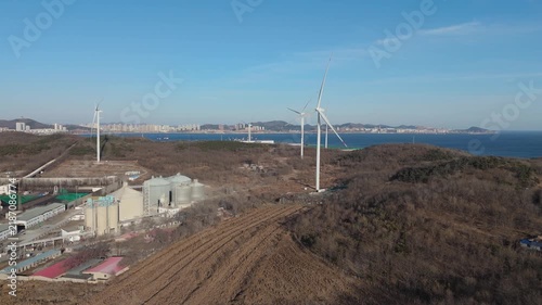 Forward-moving aerial dolly shot approaching wind turbines and industrial factory in a winter setting with leafless vegetation and clear atmospheric conditions.
