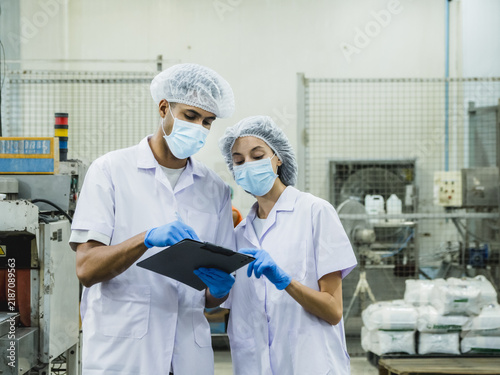 Two professional food factory inspectors in white lab coats, hairnets, surgical masks, and gloves review quality control checklists on a clipboard inside a clean manufacturing facility.