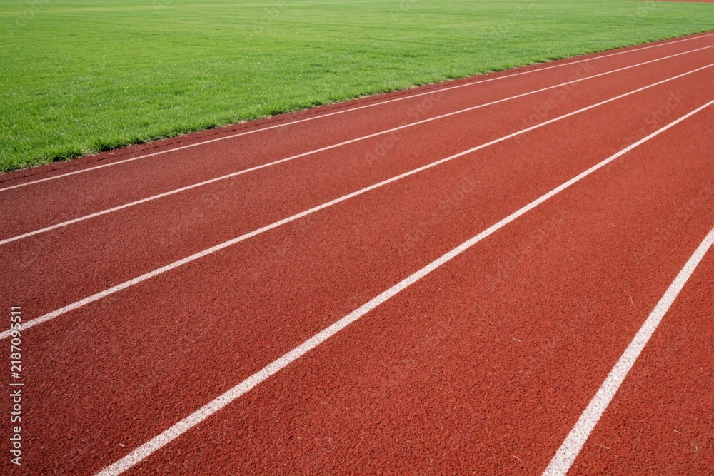 Obraz premium Red running track with white lines next to green grass field. Athletic stadium surface for sports competition, training. Outdoor arena background for sprint race, marathon, fitness lifestyle. Empty