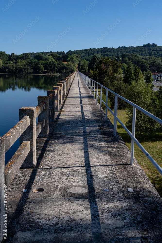 Obraz premium Narrow concrete pedestrian bridge pathway over calm lake leading towards green hills