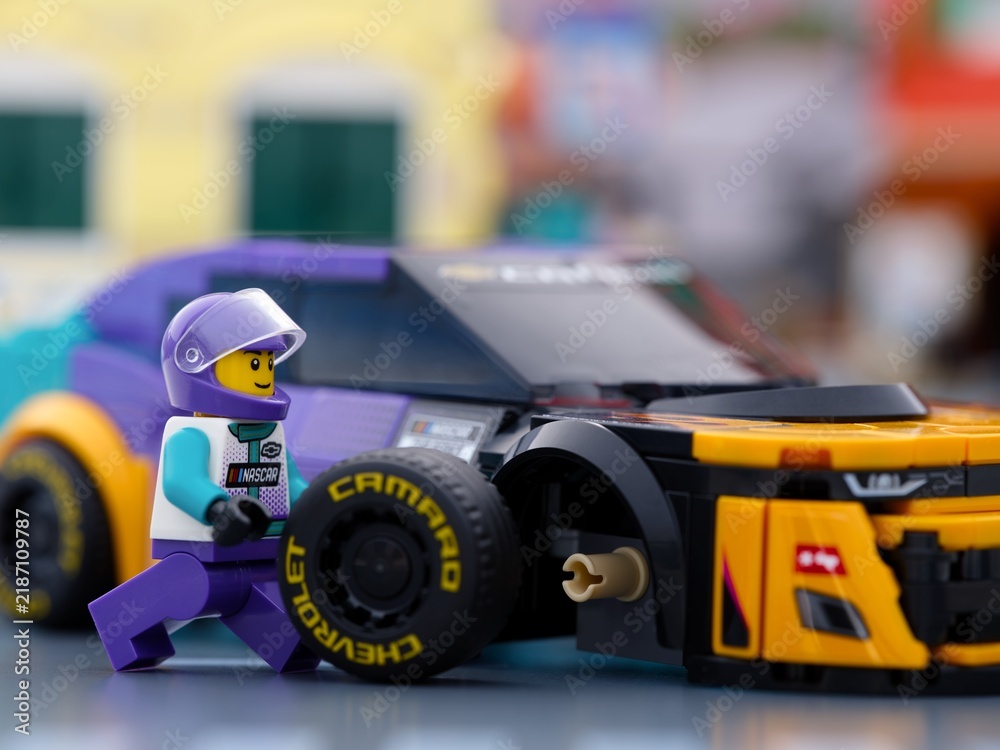 Fototapeta premium Tambov, Russian Federation - January 02, 2026 Lego NASCAR driver minifigure next to a purple and yellow Chevrolet Camaro race car during a pit stop with a detached wheel.