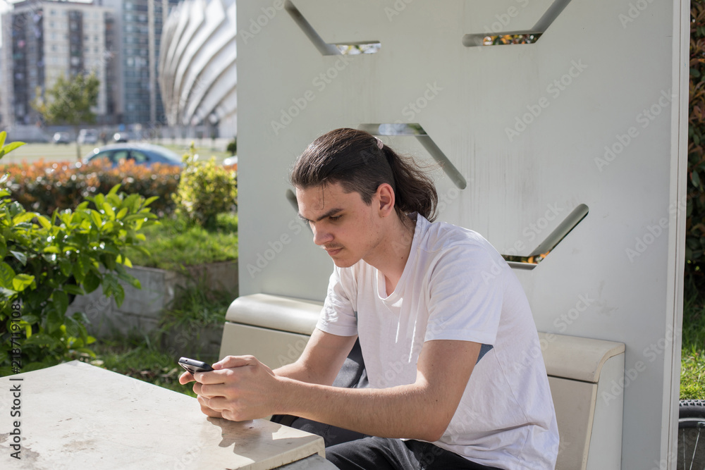 Fototapeta premium A young man in white t-shirt sits on a bench at a bus stop in an urban park. He is focused on his phone while a bicycle is parked next to him
