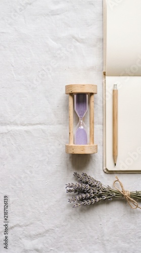 A top-down view of an hourglass with purple sand, a bundle of dried lavender, an open notebook, and a wooden pen on a light linen fabric.