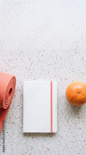 A coral yoga mat, white notebook, and grapefruit arranged on a light speckled surface, representing health and wellness.