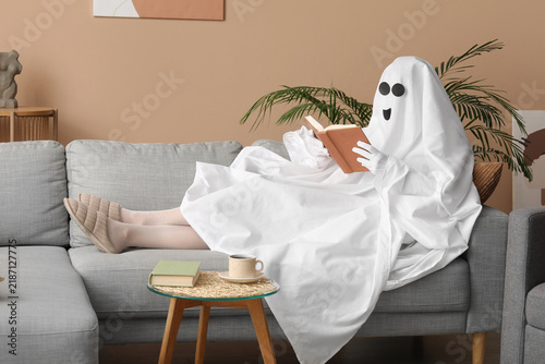 Person in a ghost costume reading a book on a gray sofa in a cozy living room, halloween and relaxation concept, a small side table with a coffee cup and another book is visible next to the sofa