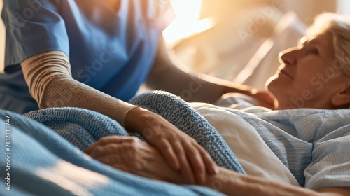Healthcare professional in blue scrubs gently comforts elderly female patient in hospital bed with soft blanket and warm lighting illuminating the room