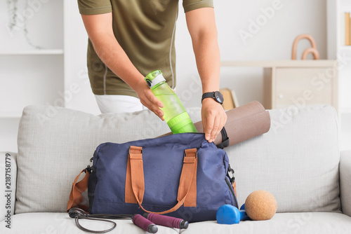 Man putting bottle of water into sports bag on sofa at home