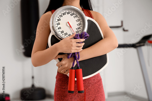 Sporty young woman with skipping rope and scales in gym. Closeup