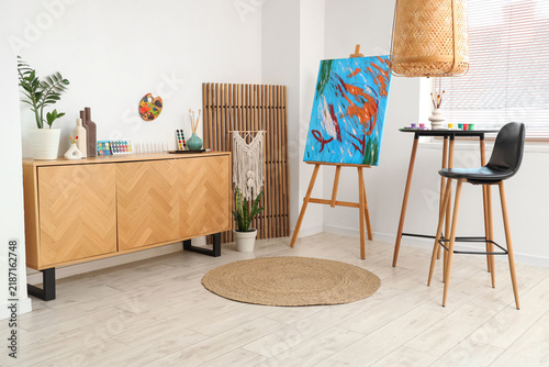 Picture on easel, chest of drawers and table with chair in painters workshop