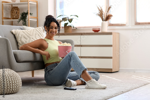 Beautiful young African-American woman with bucket of tasty popcorn sitting o...