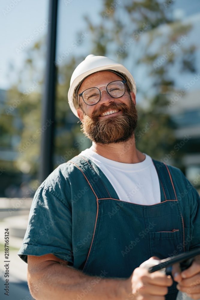 Fototapeta premium Smiling bearded construction worker wearing a white helmet and overalls outdoors for builder profile and industrial portrait banner