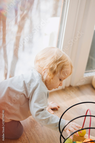 Curious toddler playing with a colorful wooden bead maze toy while sitting on the floor at home. Close-up candid lifestyle image of early childhood development, learning through play 