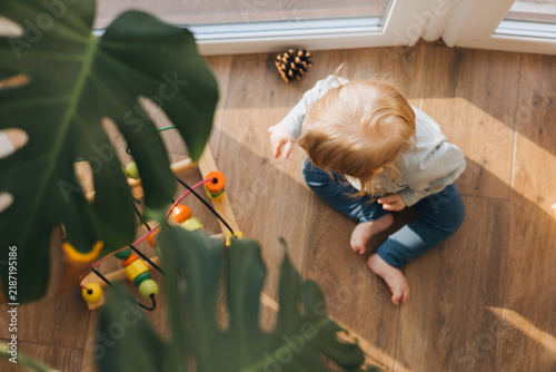 Little toddler sitting barefoot on a wooden floor and playing with a colorful educational toy near a large houseplant. Candid everyday childhood moment in a bright home with natural sunlight.
